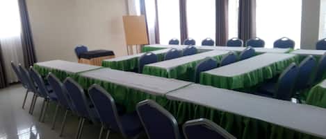 Un salón de conferencias con varias mesas y sillas, un ventilador de techo y ventanas grandes con cortinas.