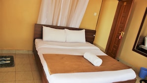 Cama bem arrumada com lençóis brancos e castanhos, cabeceira de madeira e um cesto azul no chão.