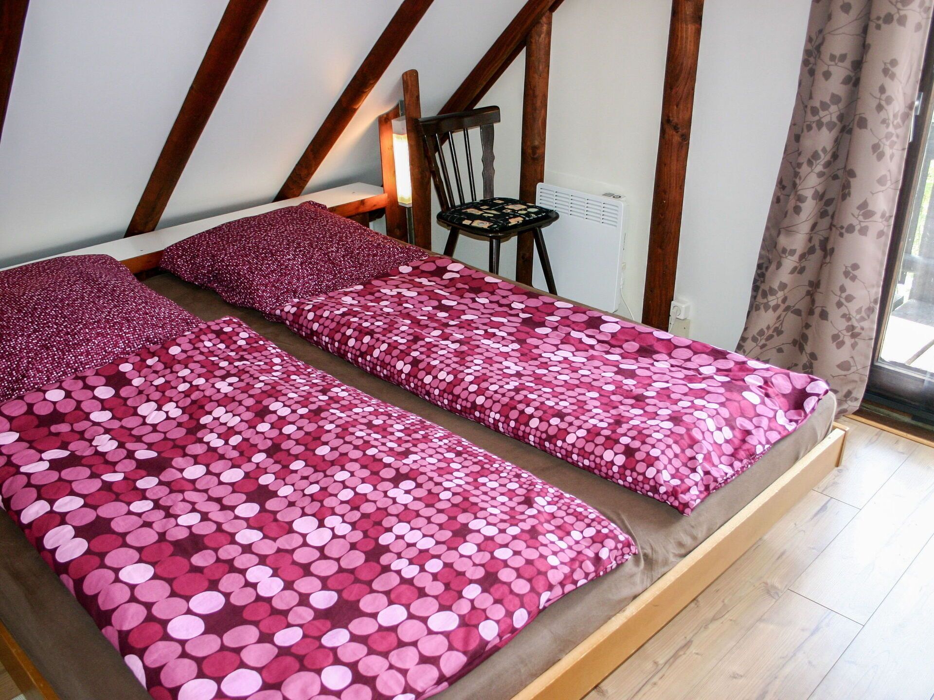 A bedroom with a wooden sloped ceiling, a bed with pink polka dot bedding, a chair, and a window with curtains.