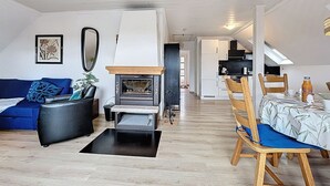 A modern living room with a fireplace, a blue sofa, a dining table with chairs, and a kitchen in the background.