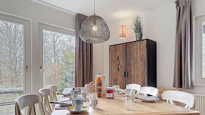 Een eethoek met een houten tafel, witte stoelen, een hanglamp en een kast met een plant erboven.