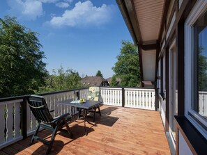 Um deck de madeira com mesa e cadeiras, um corrimão branco e vista para casas e árvores ao fundo.