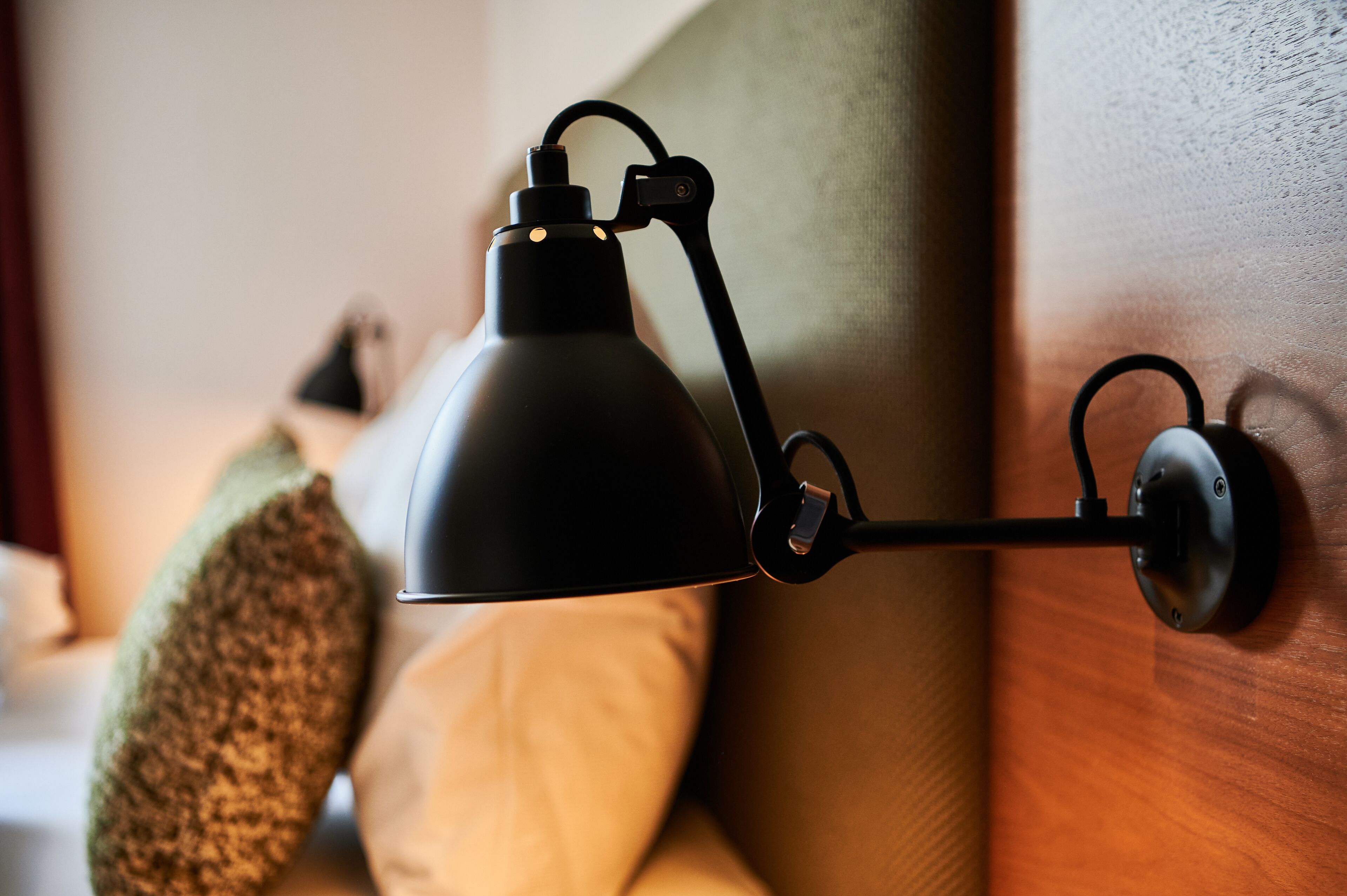 A black desk lamp attached to a wall, with a cushion and a pillow on a bed in the background.