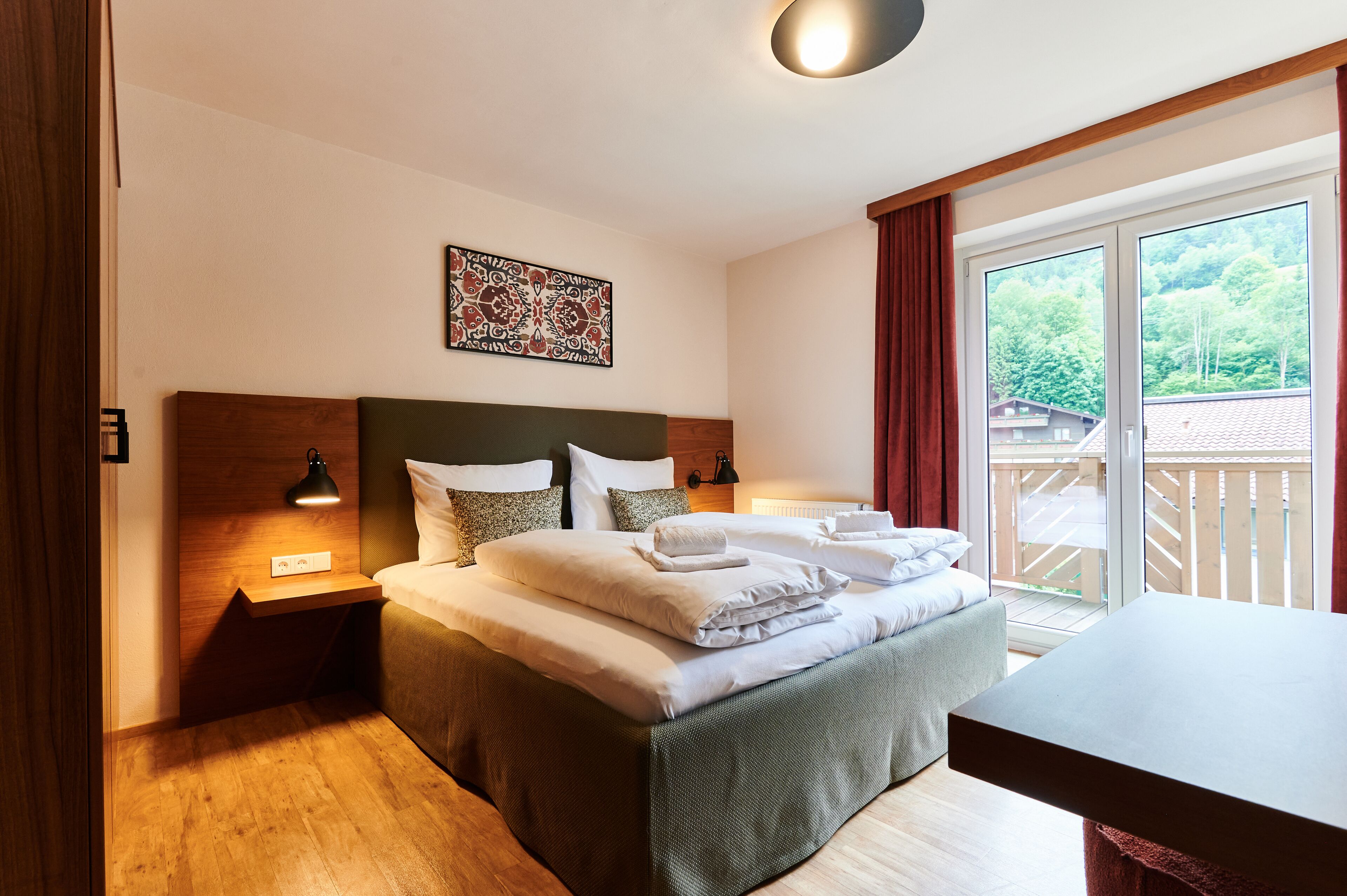 Ein ordentlich bezogenes Bett mit Kissen und Decke, ein Nachttisch mit Lampe, ein an der Wand hängendes Kunstwerk und ein großes Fenster mit Blick auf Grünflächen und ein Gebäude.