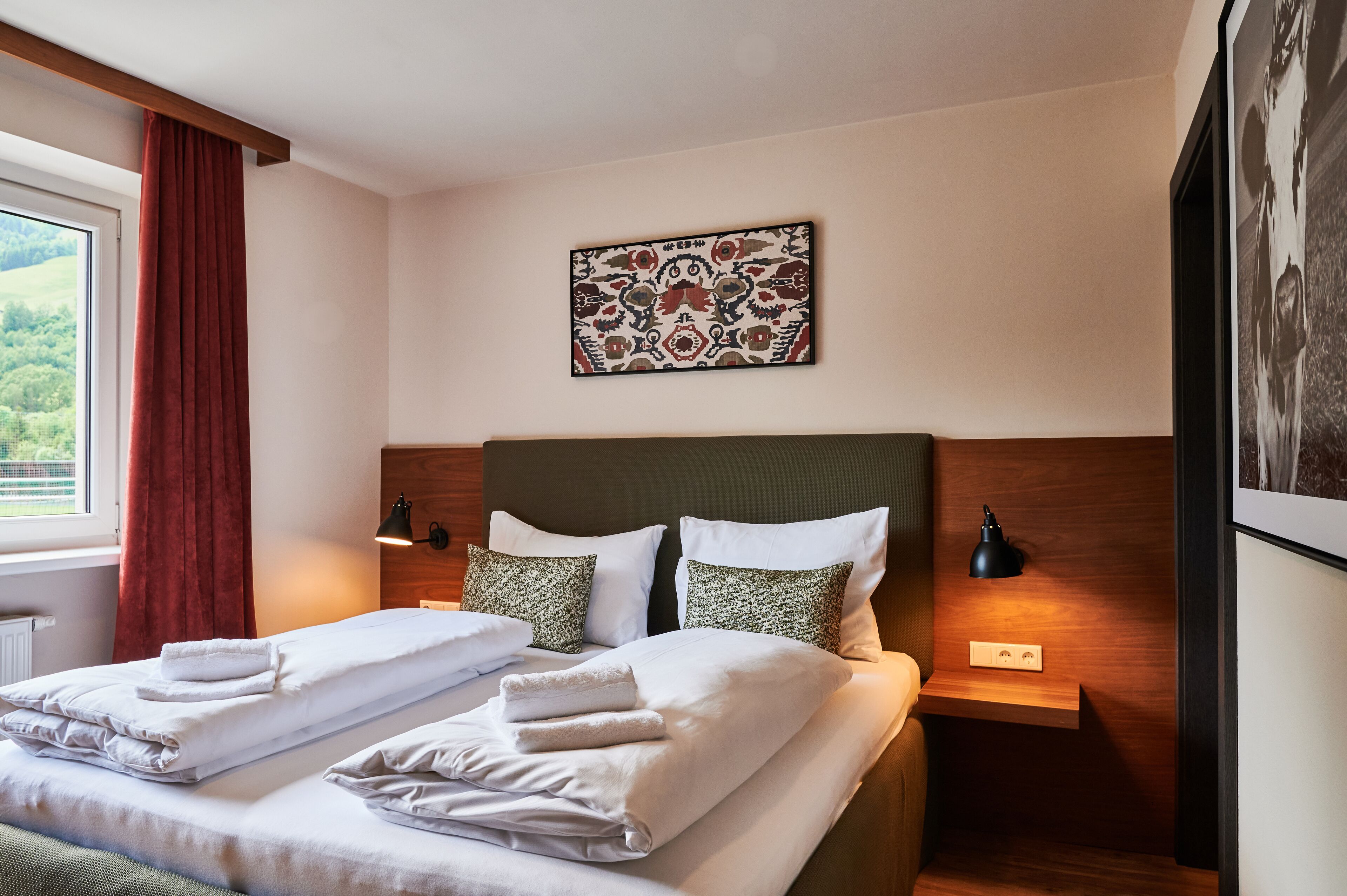Un lit double bien fait, avec du linge de lit et des serviettes blanches, une tête de lit ornée d’un motif, deux lampes de chevet et une œuvre d’art encadrée au-dessus du lit.