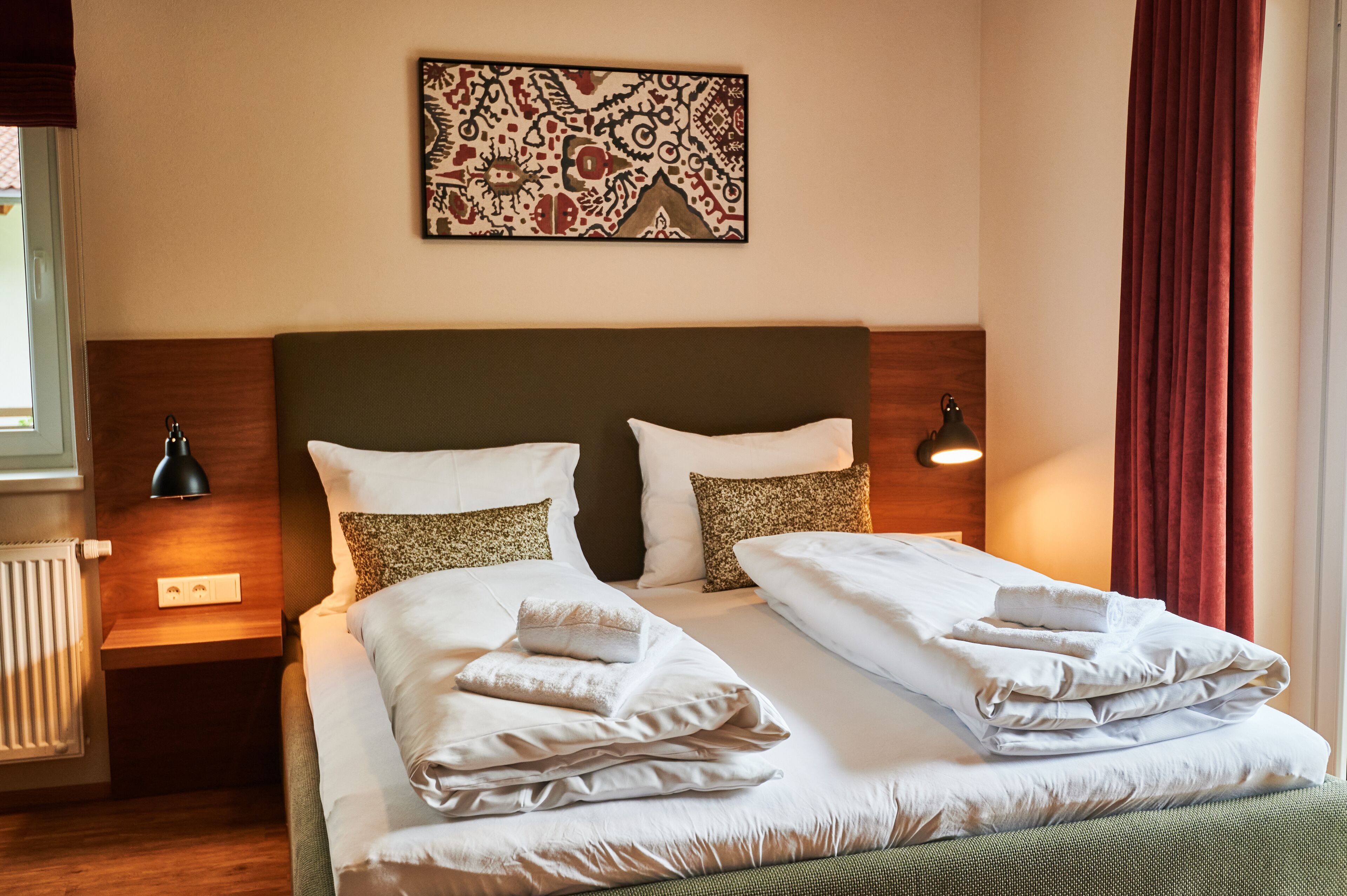 A hotel room with a double bed, two bedside tables with lamps, a wall art piece, and a window with curtains.
