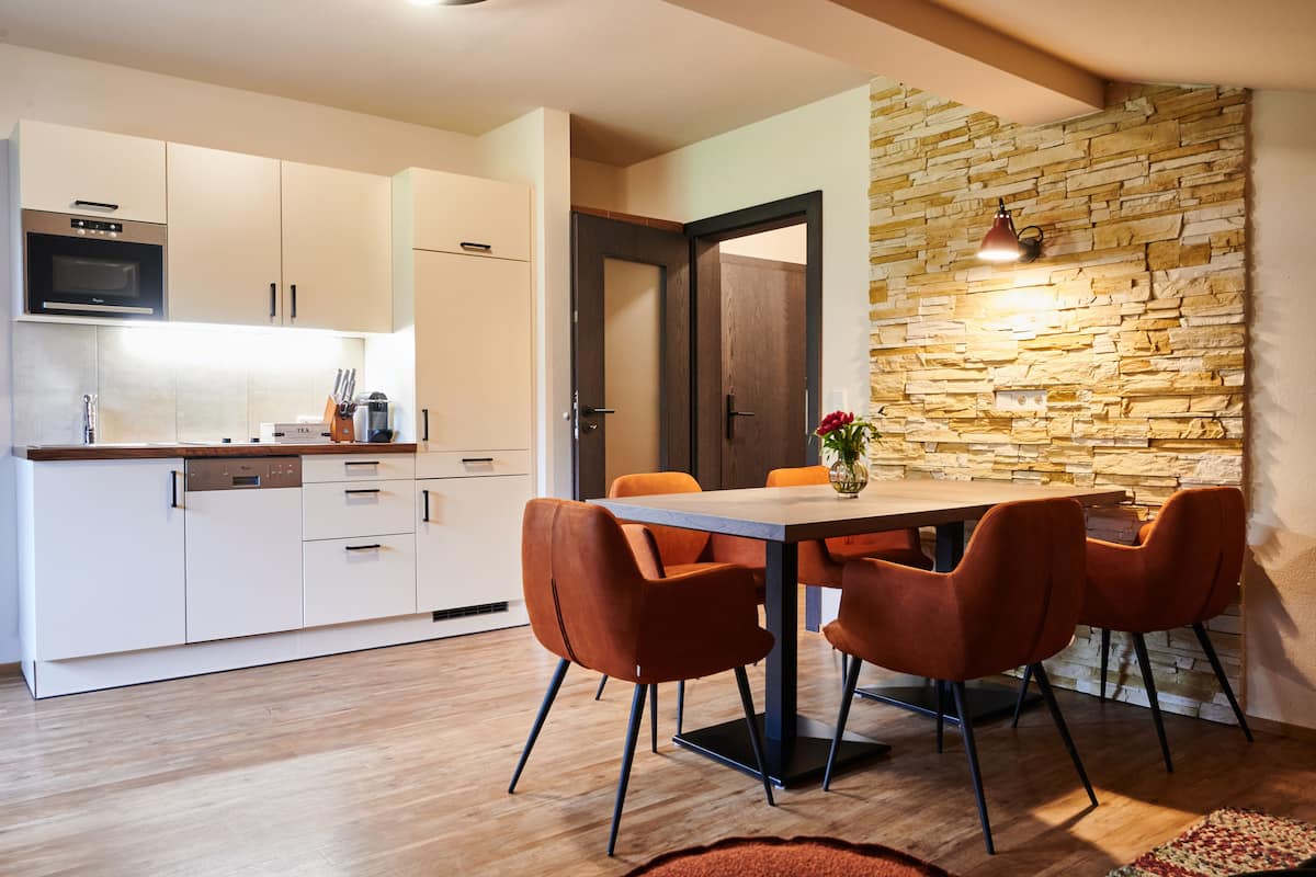 A modern kitchen with white cabinets, a built-in oven, and a dining area with a stone wall and orange chairs.