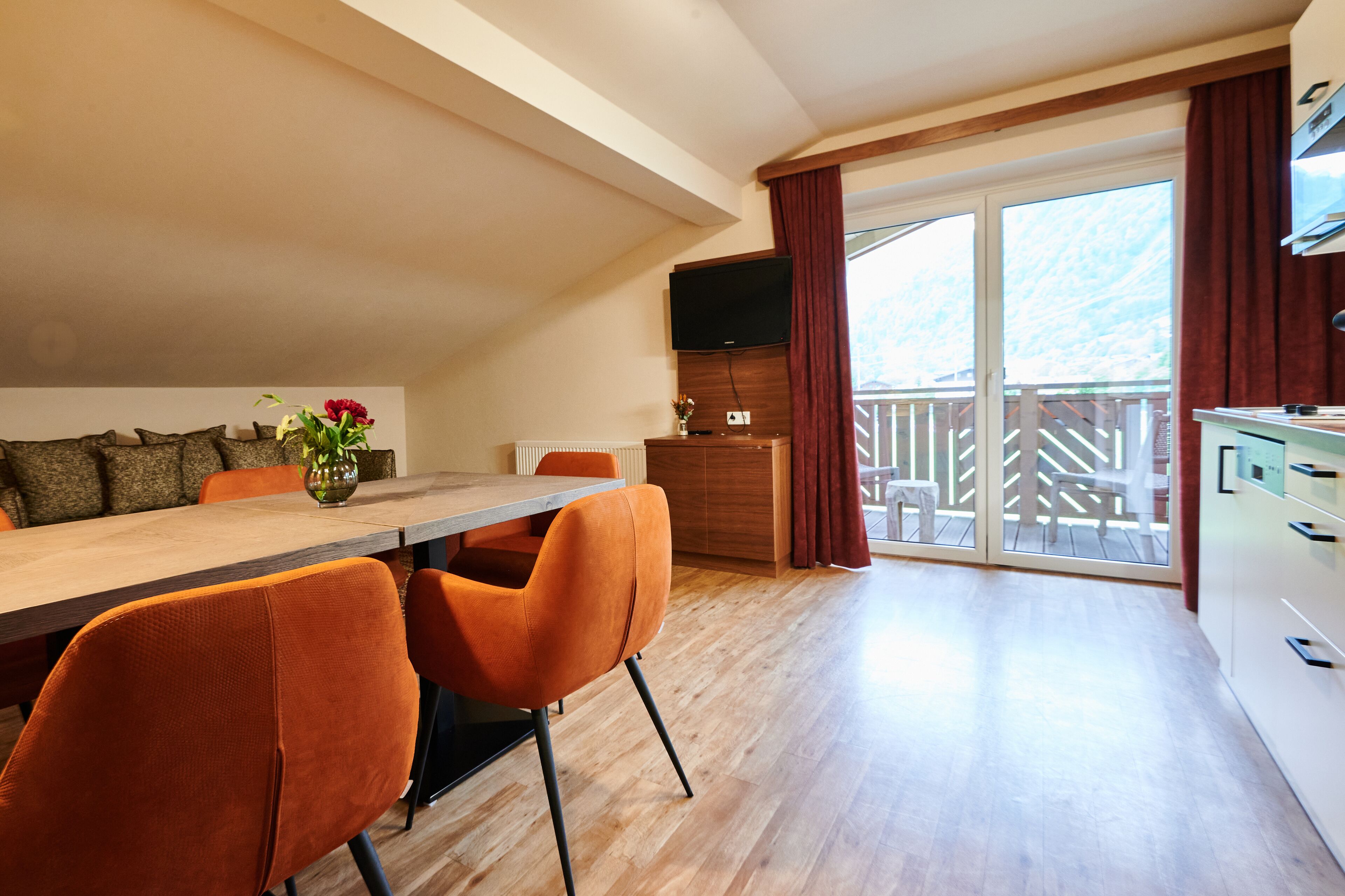 A dining area with a wooden table and orange chairs, a kitchen with a TV, and a balcony with a view of the mountains.