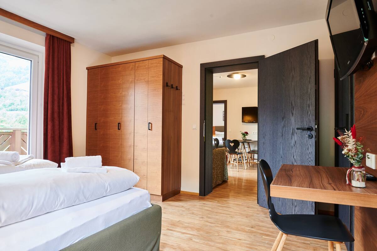 A hotel room with a bed, a wooden wardrobe, a dining table with chairs, and a view of the outdoors.