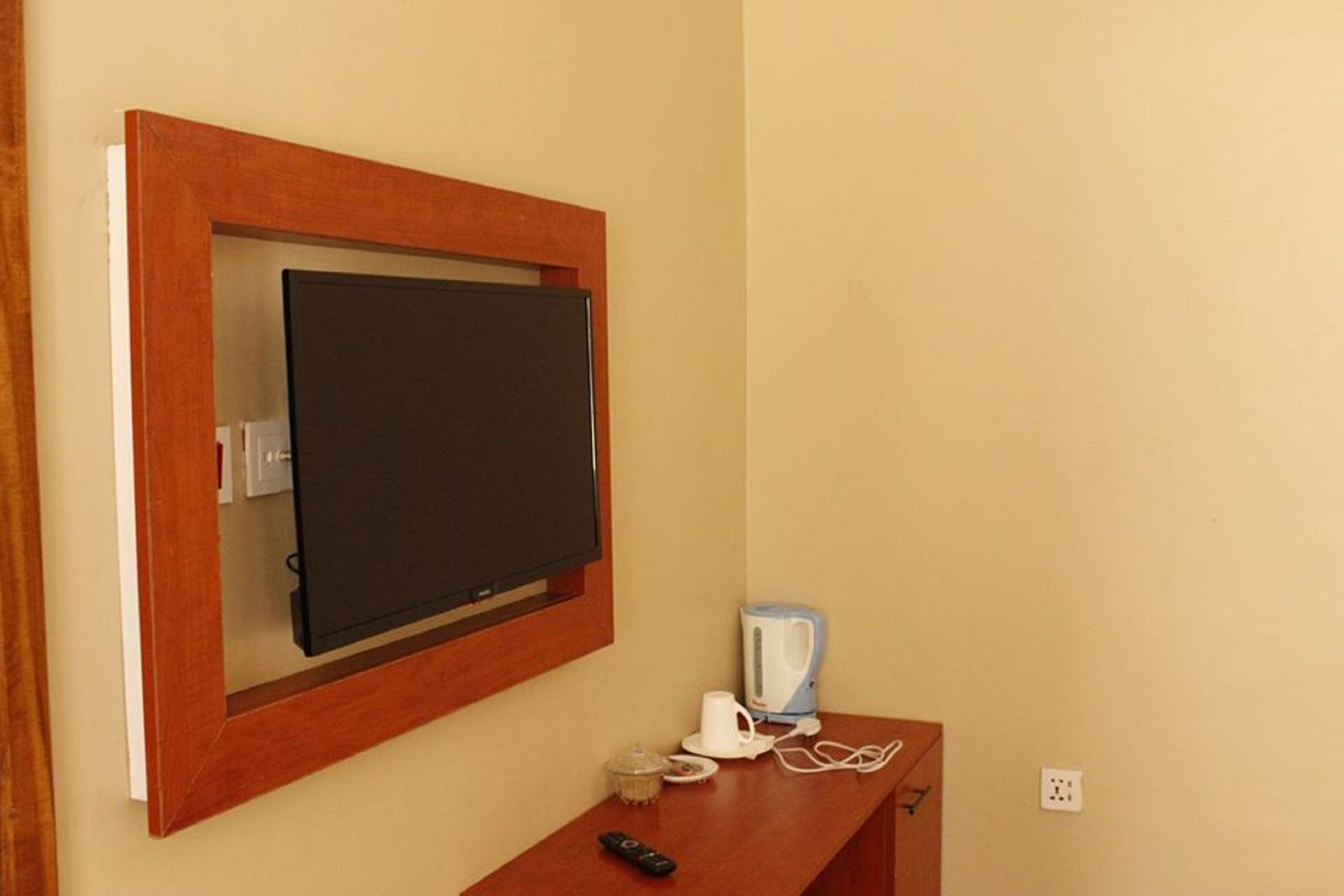 Een televisie bevestigd aan een muur, met houten lijst, een afstandsbediening, een witte koffiezetapparaat en een klein glazen vaasje op een houten tafel eronder.