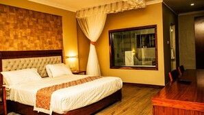 A hotel room with a large bed, a wooden headboard, a dining table, and a window with a view. - Siritamu Resort (Bungoma)