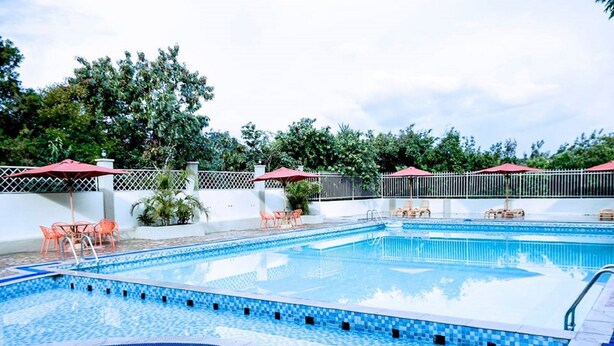 A rectangular swimming pool with blue mosaic tiles, surrounded by a white wall with a metal fence. There are orange chairs and red umbrellas around the pool area. - Siritamu Resort (Bungoma)