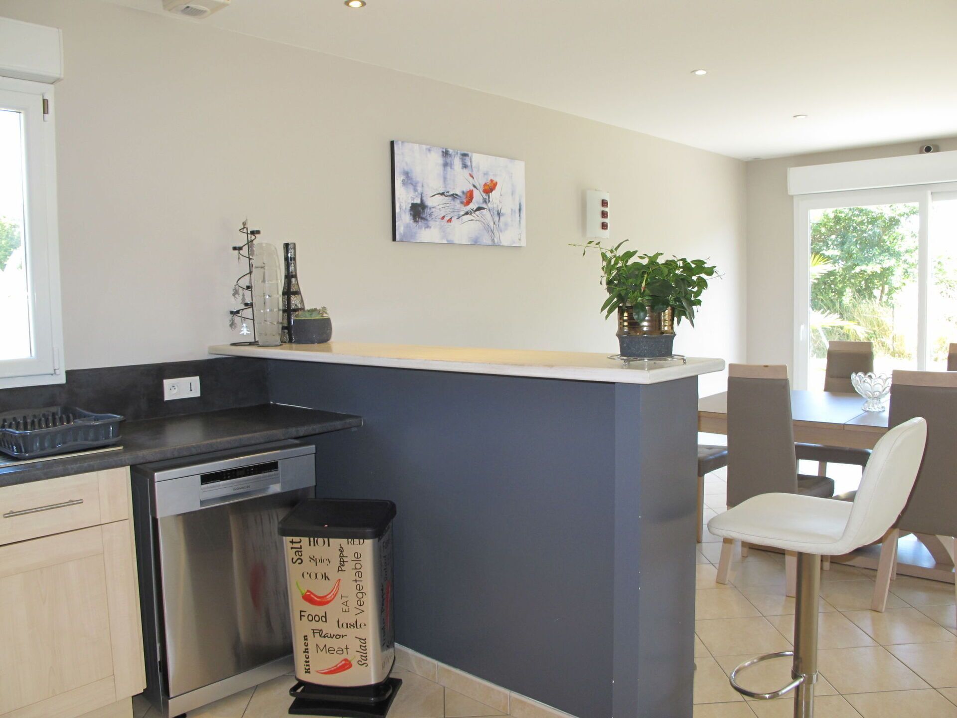 Una cocina moderna con una isla central, lavavajillas, contenedor de basura y un comedor con sillas y mesa.