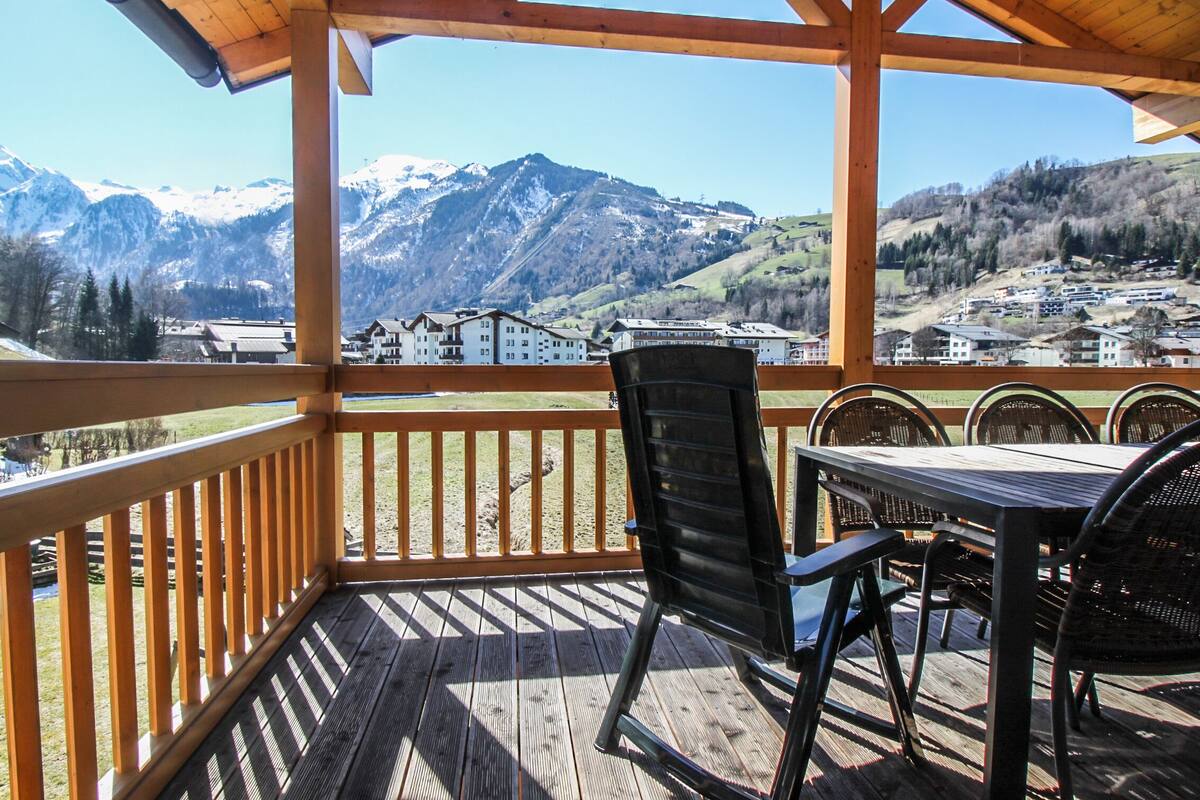 A wooden deck with a table and chairs, overlooking a mountainous landscape with buildings in the distance.