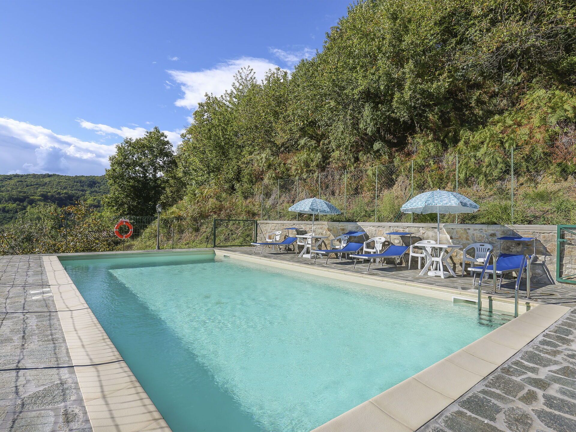 A swimming pool with lounge chairs and umbrellas, surrounded by a stone deck and lush greenery.