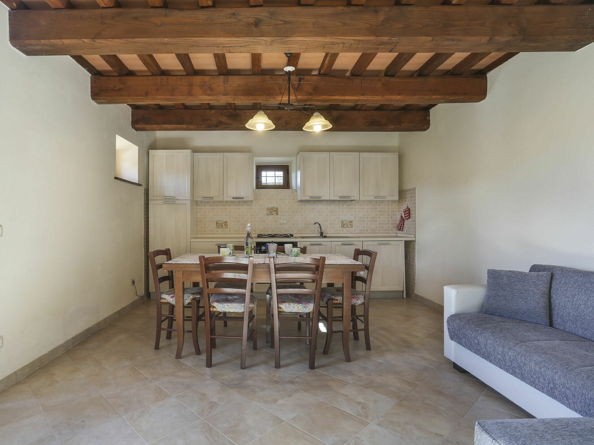 A dining area with a wooden table and chairs, a kitchen with light-colored cabinets, and a gray sofa.