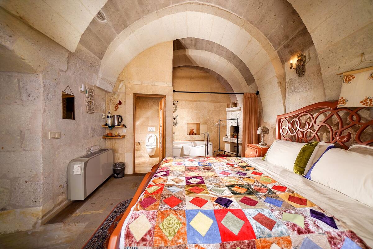 Une chambre avec un grand lit, un plafond en pierre en forme d’arc, une tête de lit en bois et une vue sur la salle de bain.