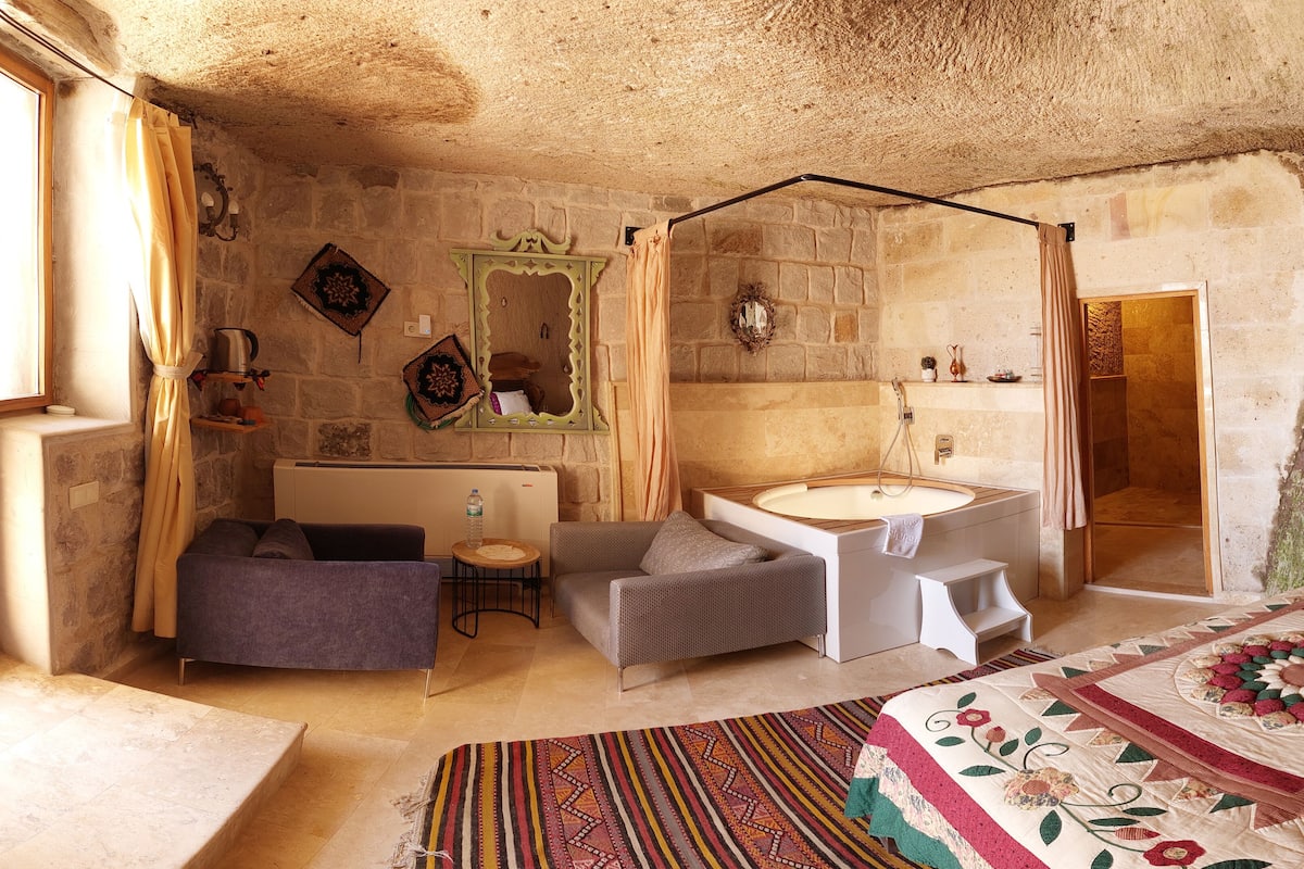 Une chambre à coucher rustique avec un mur en pierre, un lit recouvert d’une courtepointe à motifs floraux, un coin salon avec un fauteuil et une petite table, et une salle de bain dotée d’une baignoire et d’une douche.