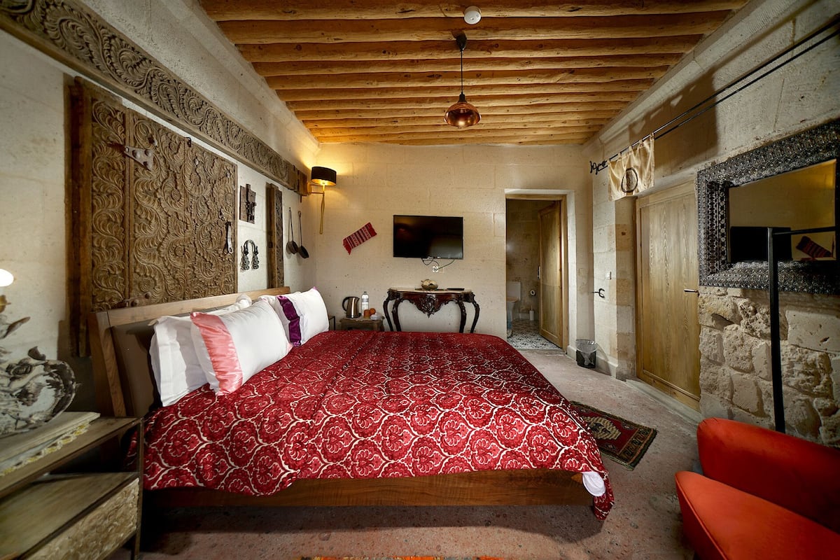 Une chambre avec un plafond en bois, un lit recouvert d’une courtepointe à motifs rouges, un téléviseur fixé au mur et un mur en pierre avec une cheminée.
