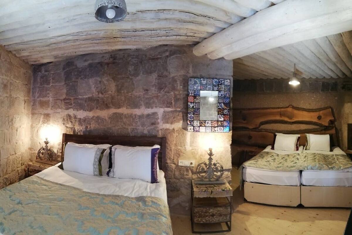 Une pièce rustique avec un mur en pierre, un plafond en bois, un lit recouvert d’une courtepointe à motifs, une table de chevet avec une lampe et une petite table ornée d’un objet décoratif.