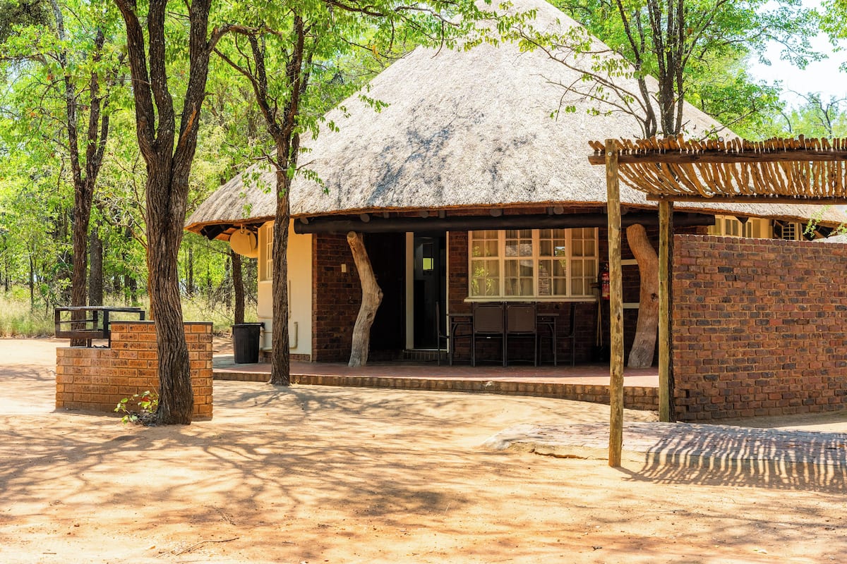 A rustic building with a thatched roof, surrounded by trees, featuring a brick wall and a wooden pergola.