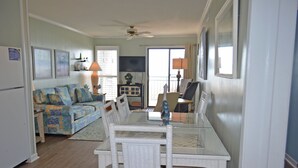A living room with a glass-top dining table, white wicker chairs, a sofa with colorful cushions, a ceiling fan, and a view through a sliding glass door. - A Place at the Beach II 2H - Ocean Front in Garden City Beach (Garden City)
