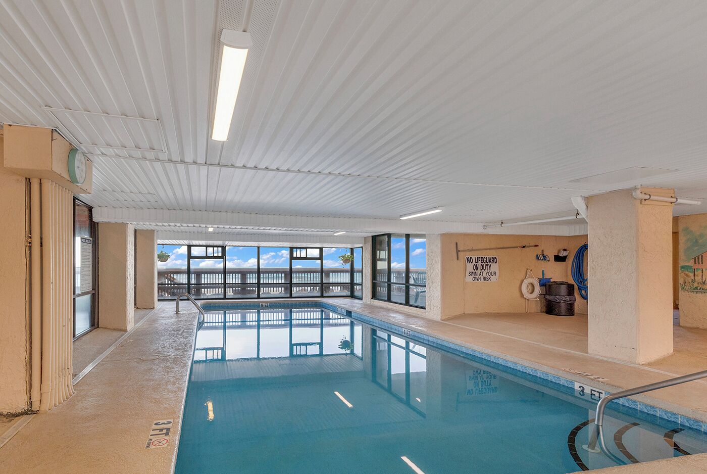 An indoor swimming pool with clear blue water, a lifeguard sign, and a view of the sky through large windows.