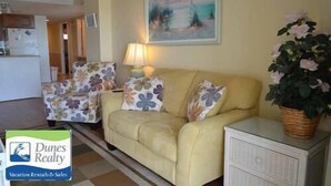 Una sala de estar con un sofá, dos sillones, una lámpara y un cuadro en la pared. Hay un cartel de Dunes Realty en la esquina inferior izquierda.