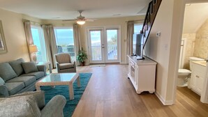 A living room with a sofa, coffee table, and TV stand. There is a balcony with curtains and a ceiling fan. - Admiral's Quarters II #10 - Oceanfront in Surfside Beach (Surfside Beach)
