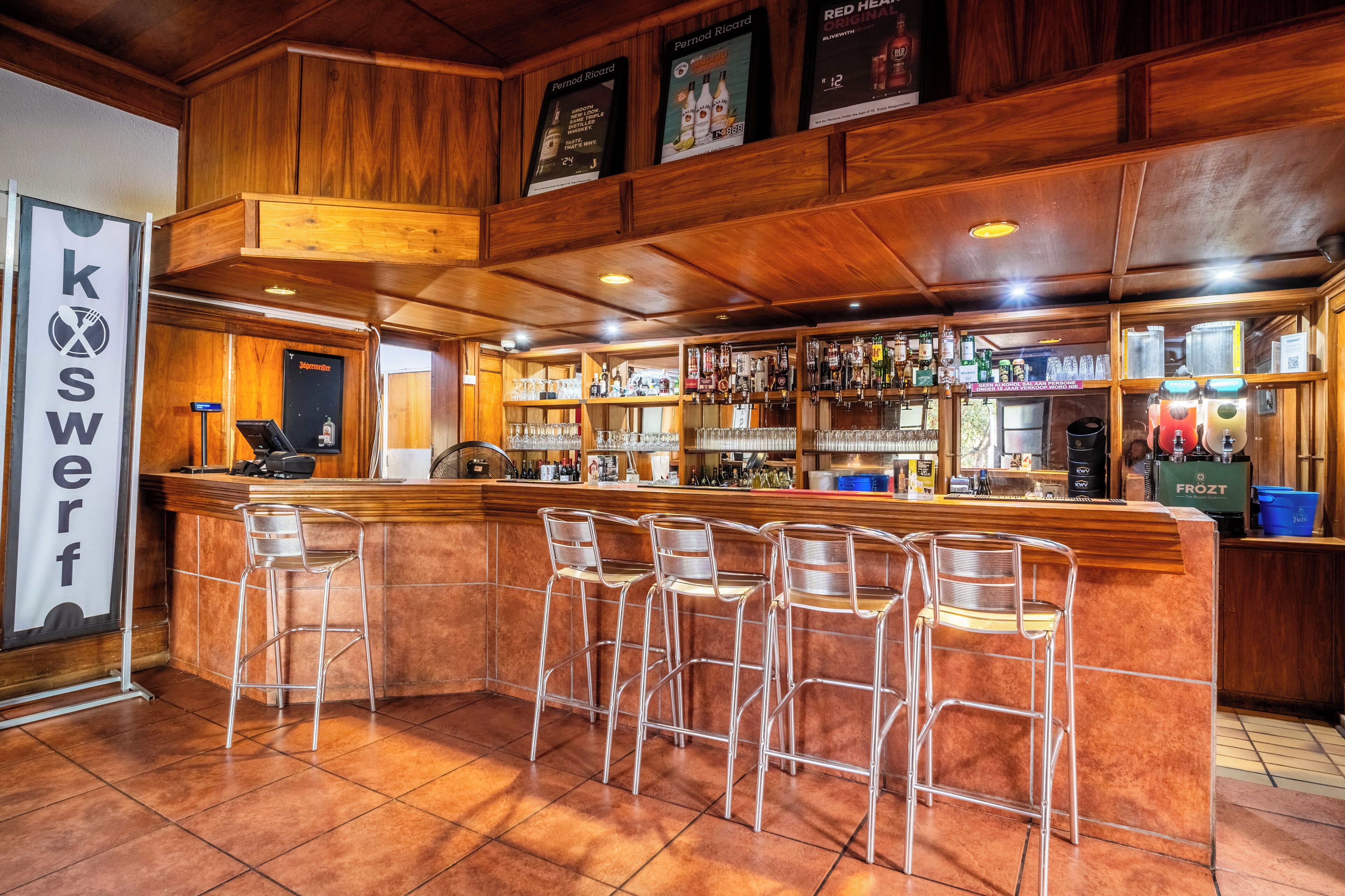 A bar area with a wooden counter, metal bar stools, and a variety of bottles and glasses on shelves.