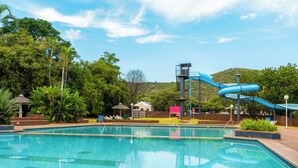 Une piscine avec un toboggan bleu, entourée de verdure et sous un ciel dégagé.