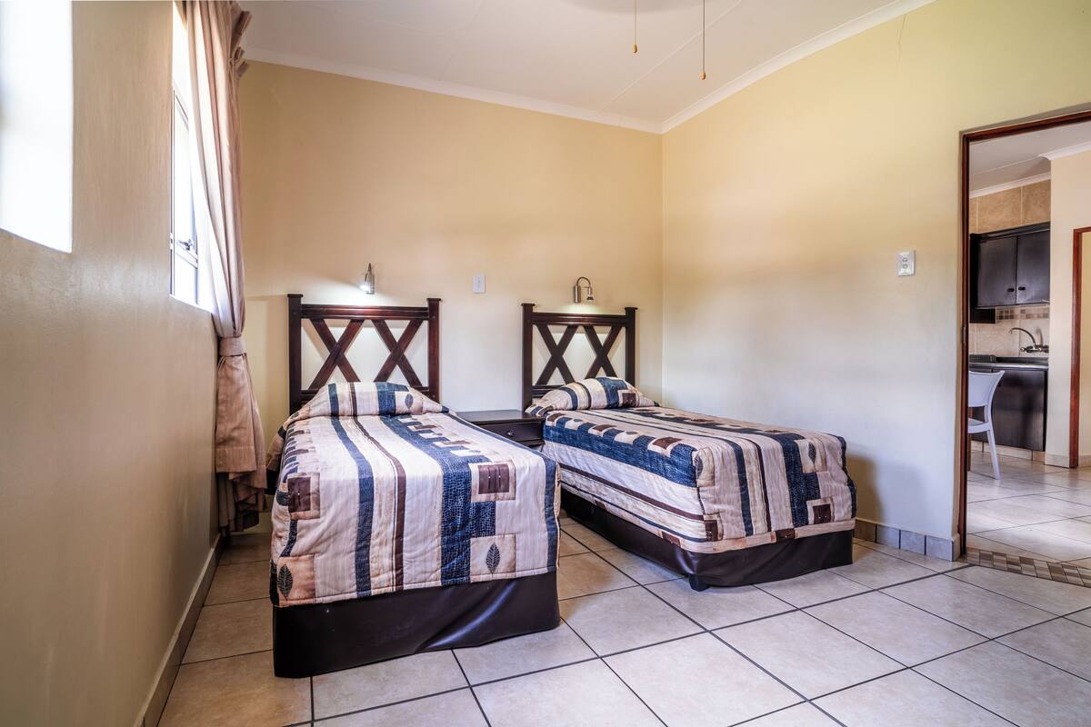 Una habitación con dos camas, una ventana con cortinas, un lavabo y vistas a una cocina.