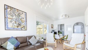 A modern living room with a brown leather sofa, white armchairs, and a white coffee table. A large abstract painting is on the wall, and there is a kitchen area in the background. - Nice apartment for 6 guests with WIFI, TV and terrace (Roses)