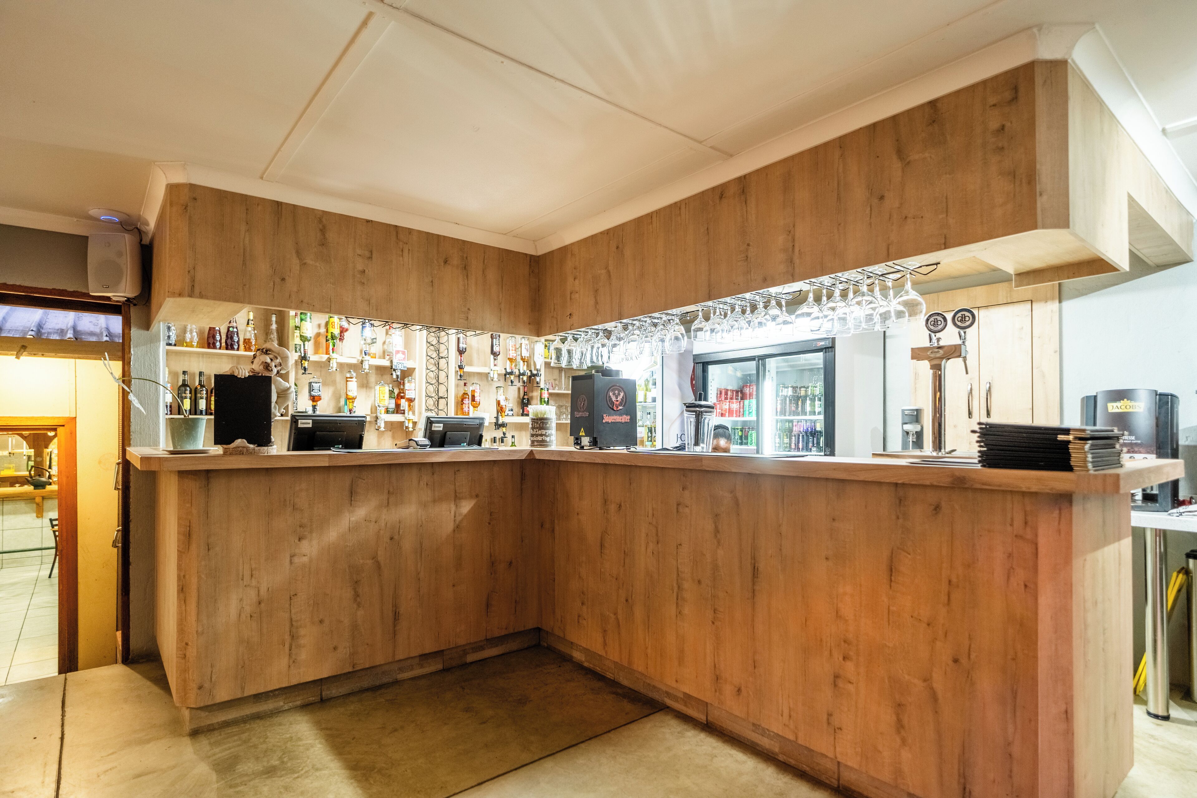 Een bar met houten lambrisering, een kassa en diverse flessen en glazen op planken.