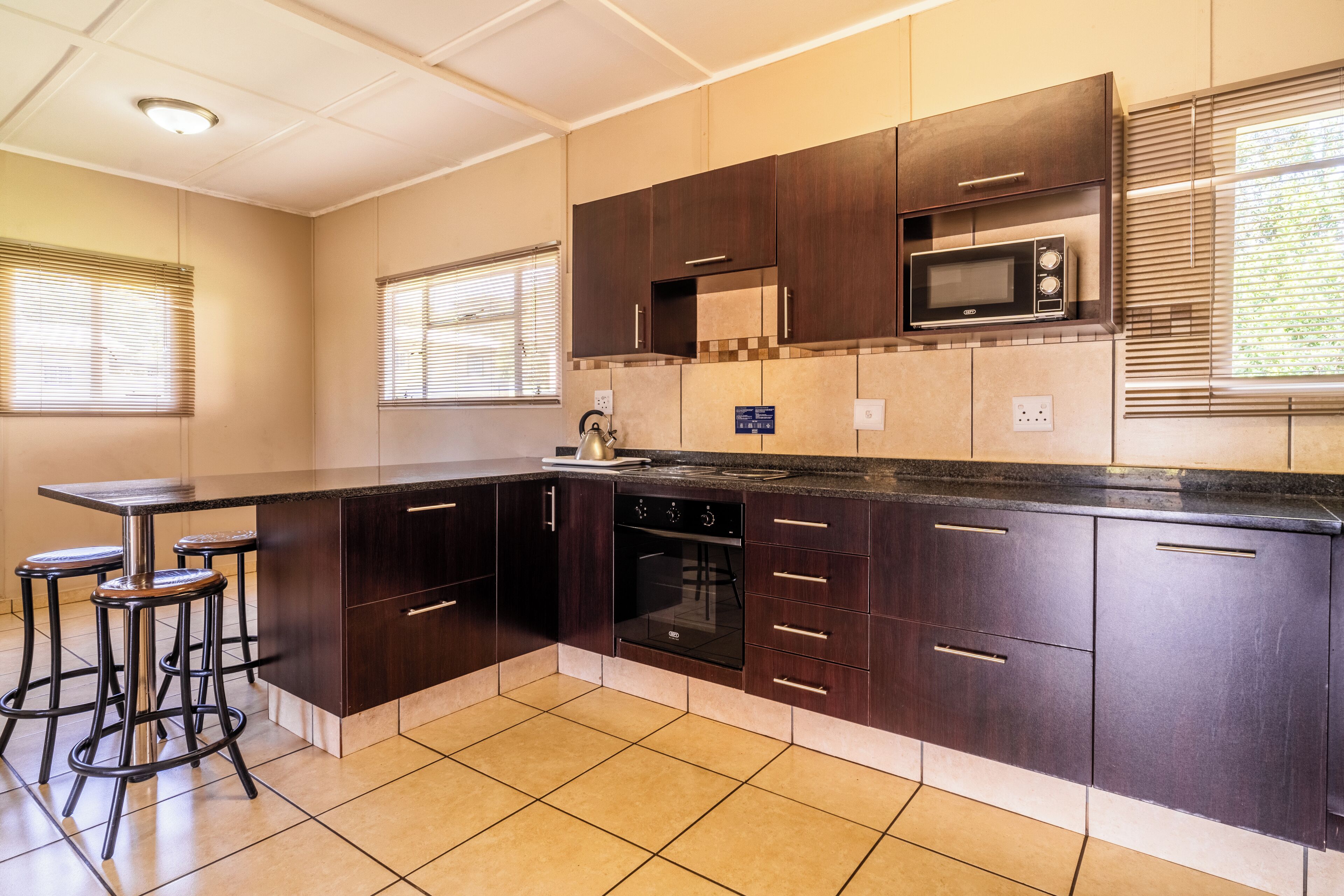 A modern kitchen with dark wooden cabinets, a black countertop, and a built-in microwave. There are three bar stools and a window with blinds.