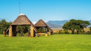 A rural setting with two thatched-roof huts with stone walls, surrounded by a grassy field and trees, with mountains in the background. - ATKV Drakensville (Jagersrust)