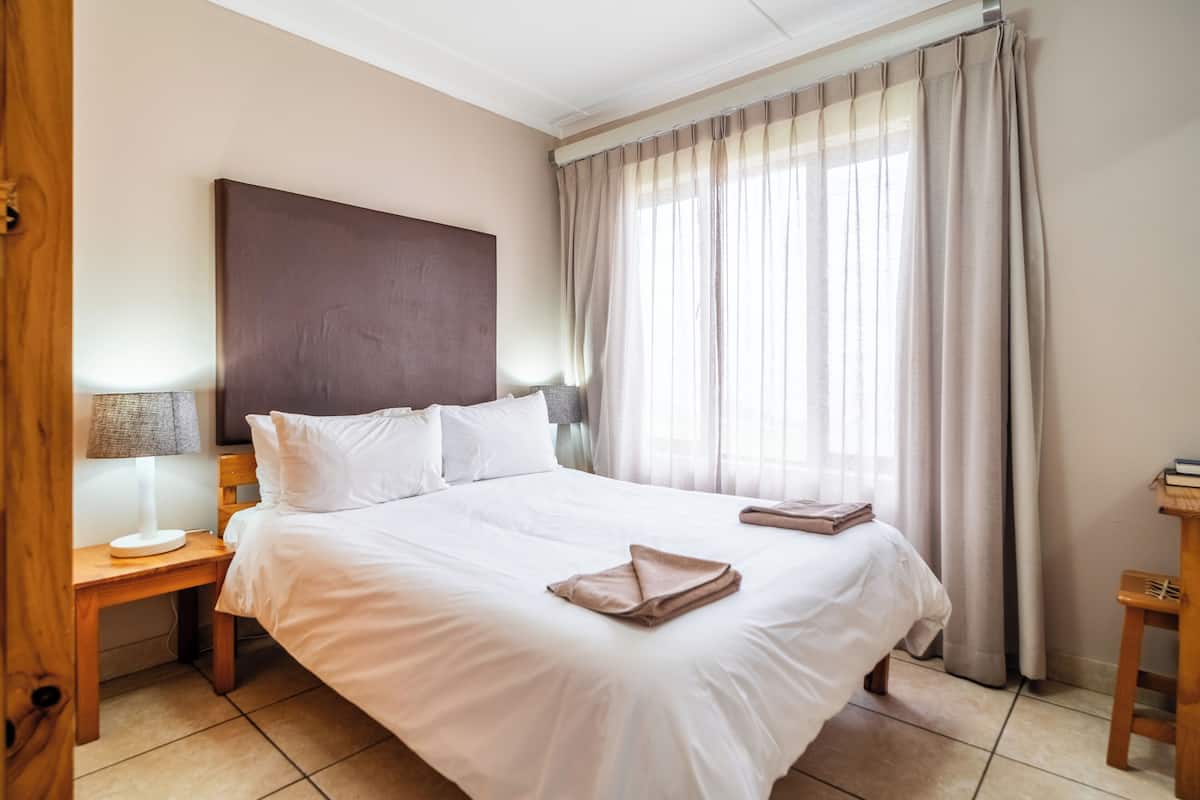 A neatly made bed with white linens and a brown towel, a bedside lamp, and a wooden nightstand.
