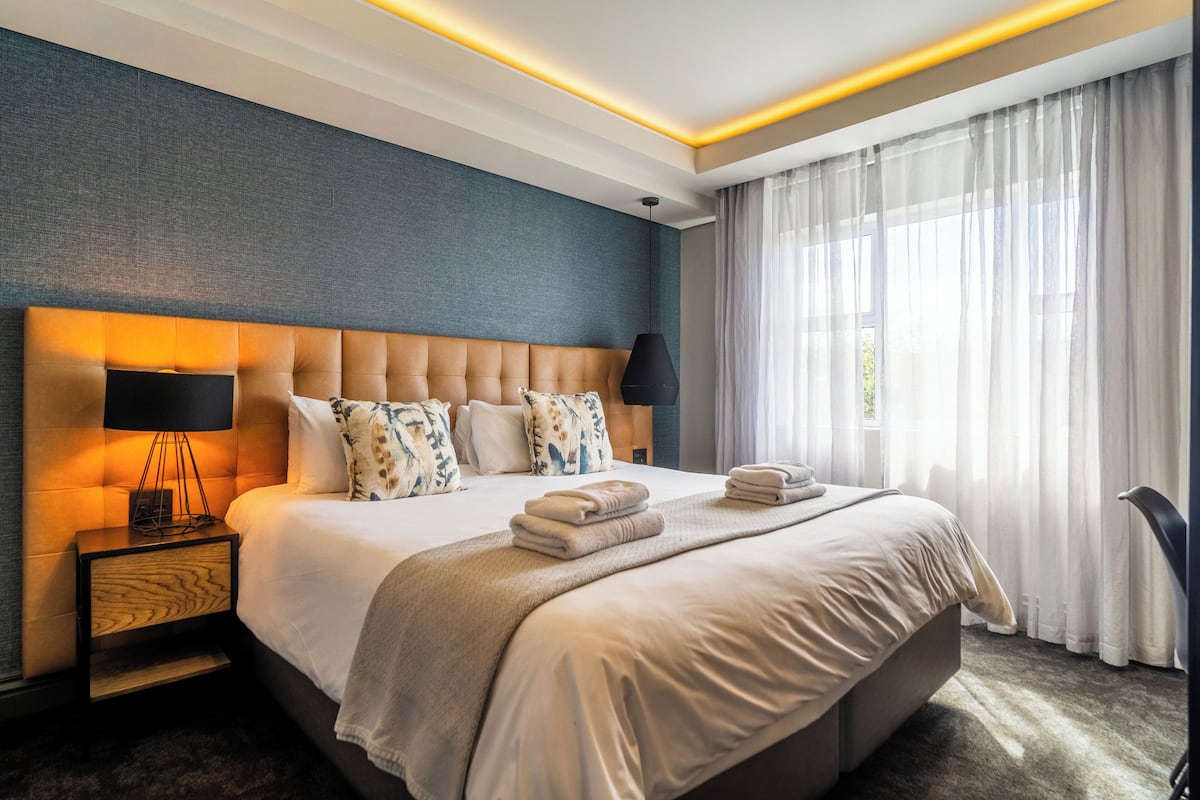 A modern hotel room with a large bed, a nightstand, a lamp, and a view of the outdoors through sheer curtains.