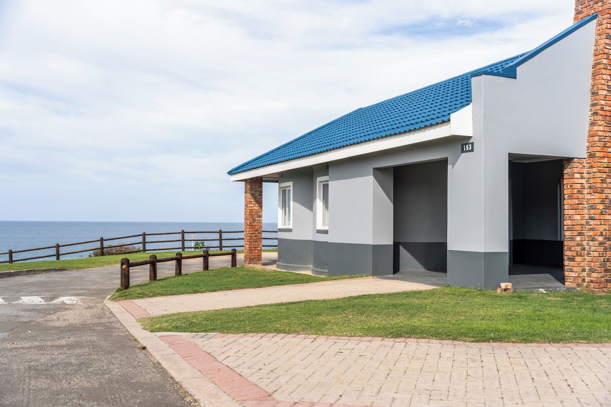 A modern building with a blue roof, brick columns, and a paved walkway leading to a wooden fence overlooking the sea.