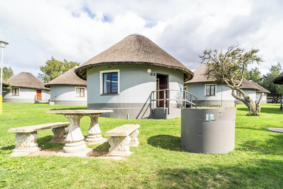 A traditional thatched-roof building with a staircase leading to the entrance, a stone bench, and a concrete water tank.