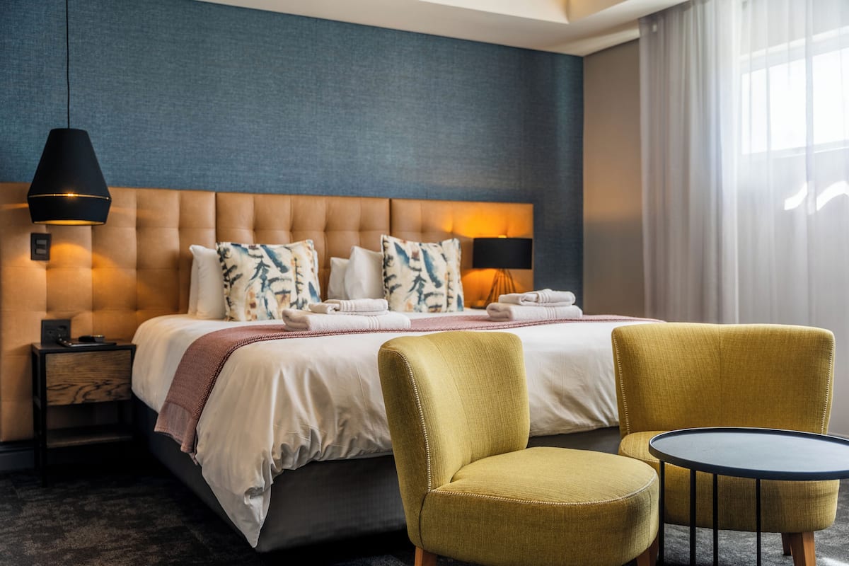 A hotel room with a large bed, two armchairs, a small round table, a nightstand, and a lamp.