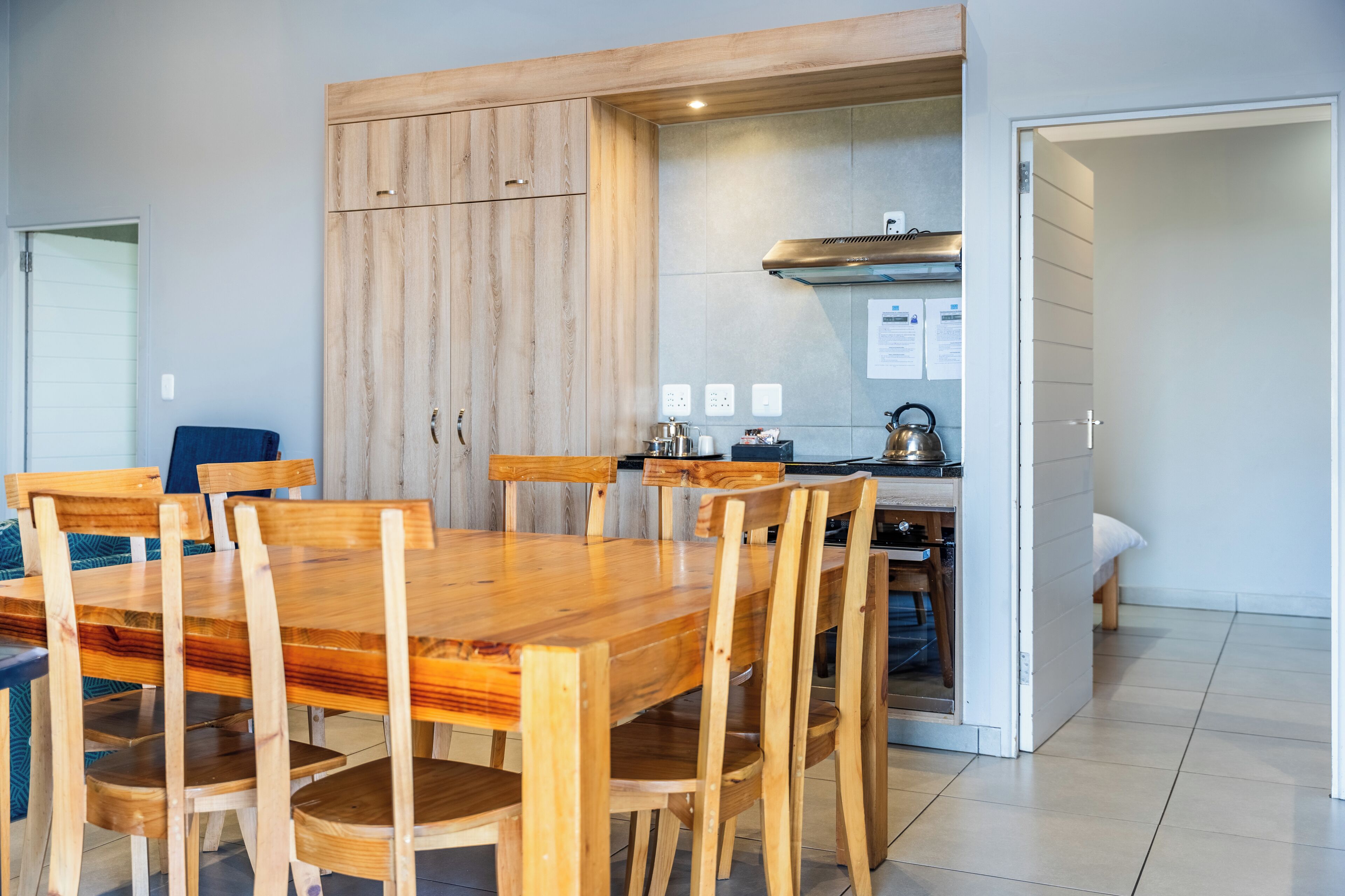 Un comedor con mesa y sillas de madera, una cocina con estufa y hervidor, y una puerta que da a otra habitación.