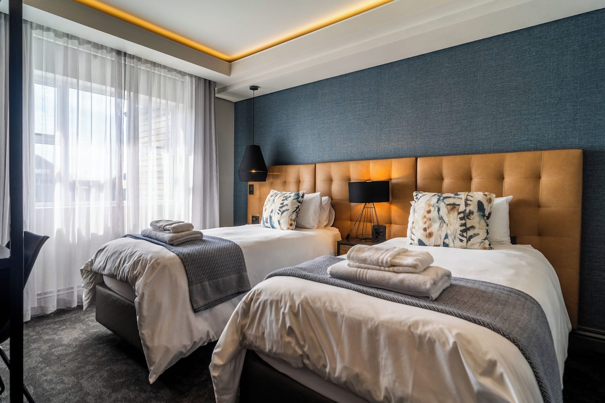 A hotel room with two beds, a large window with sheer curtains, a dark blue accent wall, and a modern chandelier.