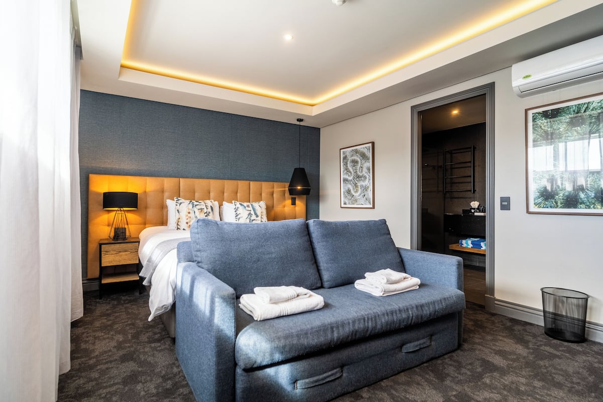 A modern hotel room with a large bed, a blue sofa, a nightstand with a lamp, and a framed artwork on the wall.