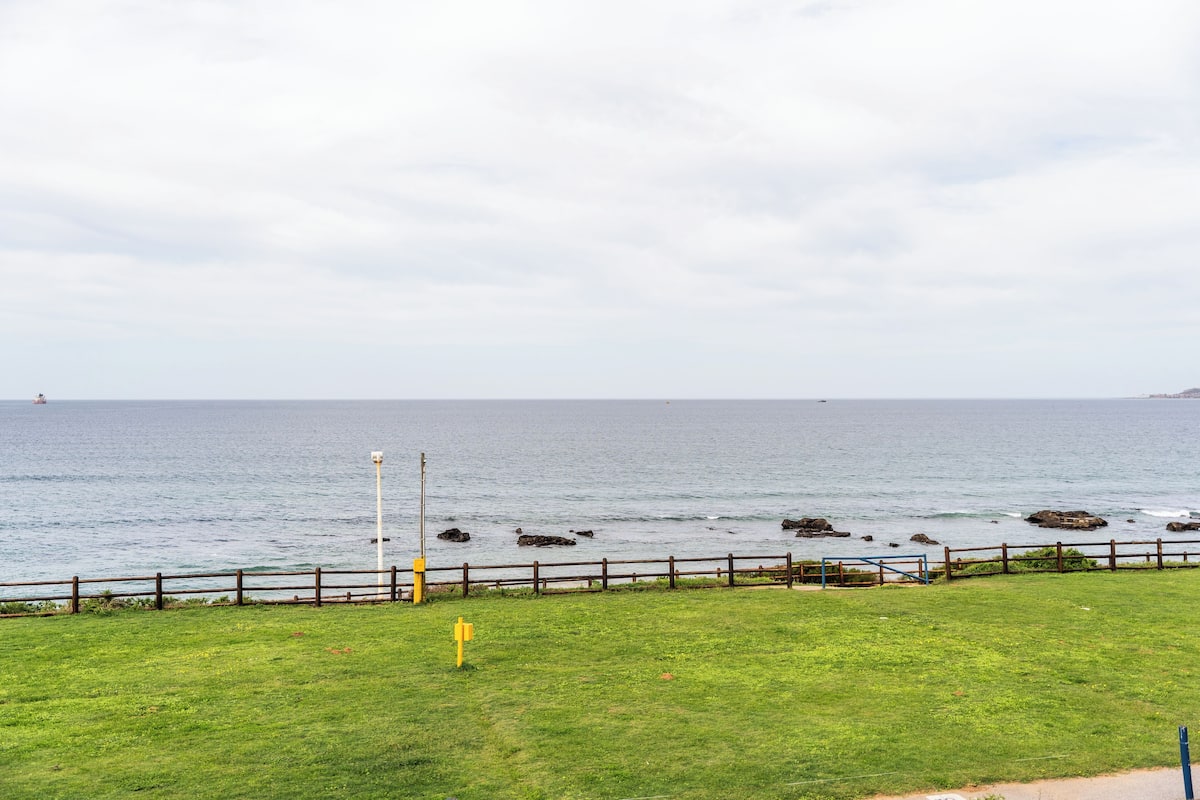 A grassy area with a wooden fence and a yellow sign, overlooking a body of water with rocks and a distant shoreline.