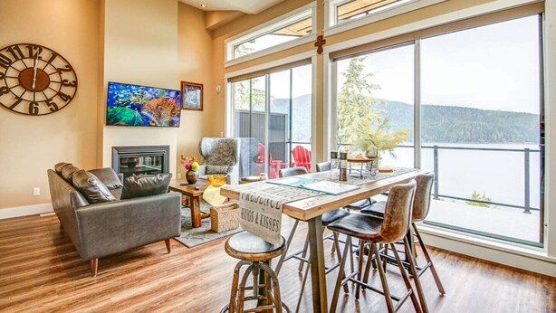 A spacious living area with a large window overlooking a body of water, a fireplace, and a dining table with chairs.