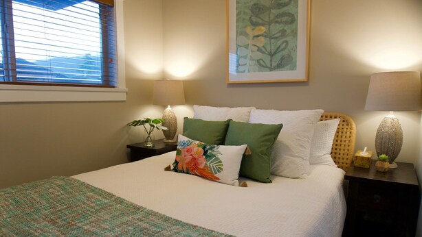 A neatly made bed with a patterned blanket, a green pillow, and a floral pillow. There is a bedside table with a lamp, a vase with a plant, and a framed picture on the wall.