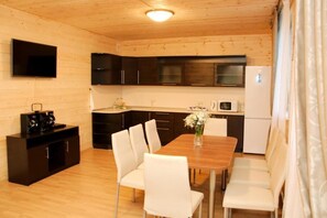 Una cocina compacta con armarios oscuros, una mesa de comedor de madera, sillas blancas, un televisor de pantalla plana montado en la pared y un refrigerador.