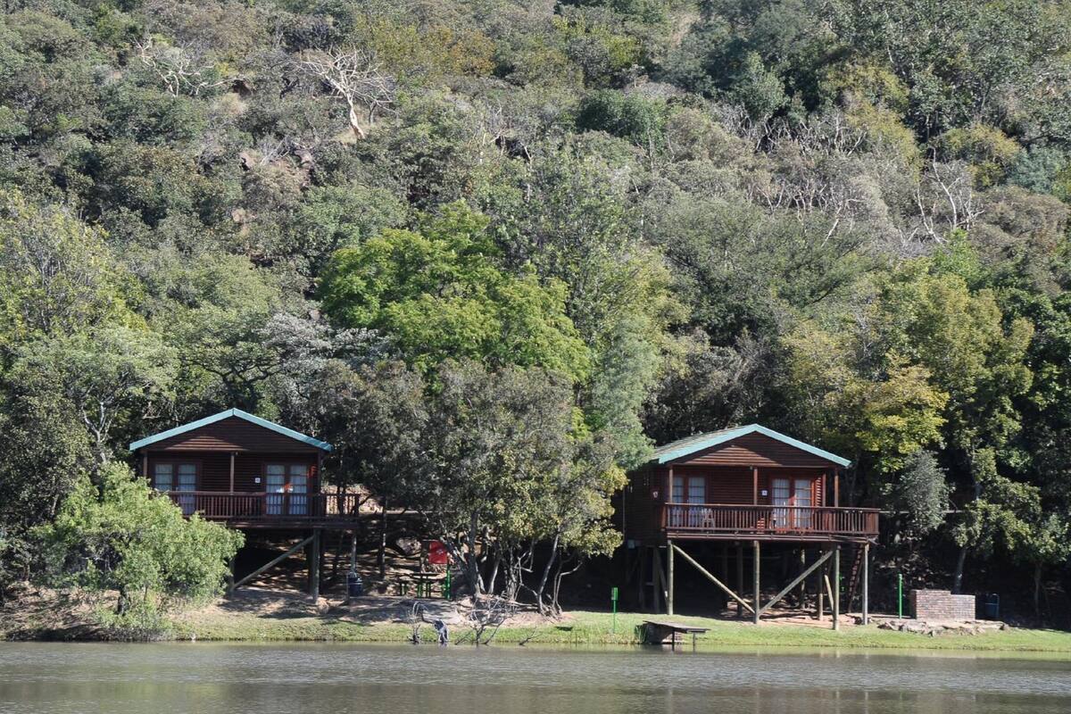Two wooden cabins with balconies, situated by a calm lake, surrounded by dense forest.