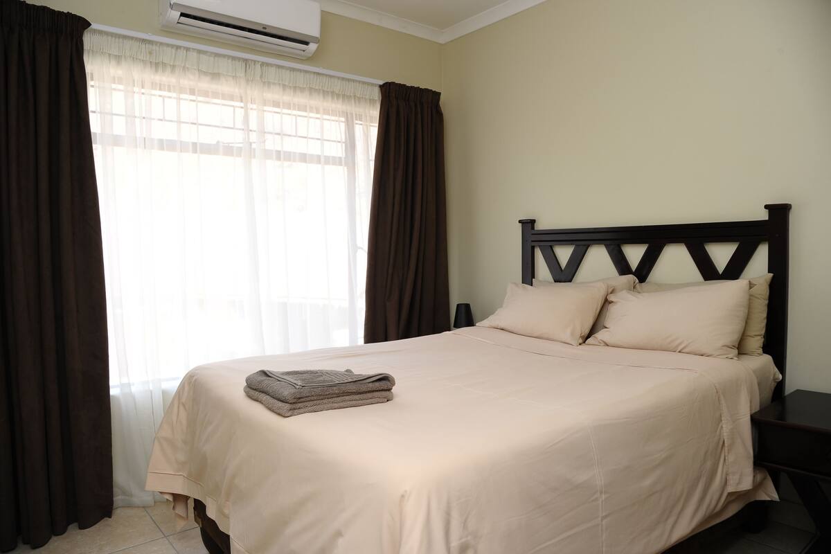 A neatly made bed with a beige bedspread and pillows, a bedside table, and a window with curtains.